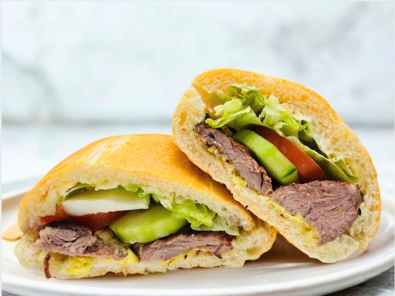Two halves of a steak sandwich with lettuce, tomato, and avocado on a white plate.